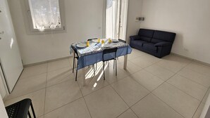 Een kamer met een eettafel voor vier personen, een blauwe bank en twee ramen.