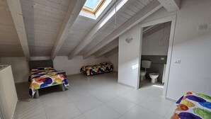Een ruime kamer onder de dakpannen met dakraam, twee bedden met kleurrijk beddengoed, een badkamer met toilet en bidet, en een vloer van witte tegels.
