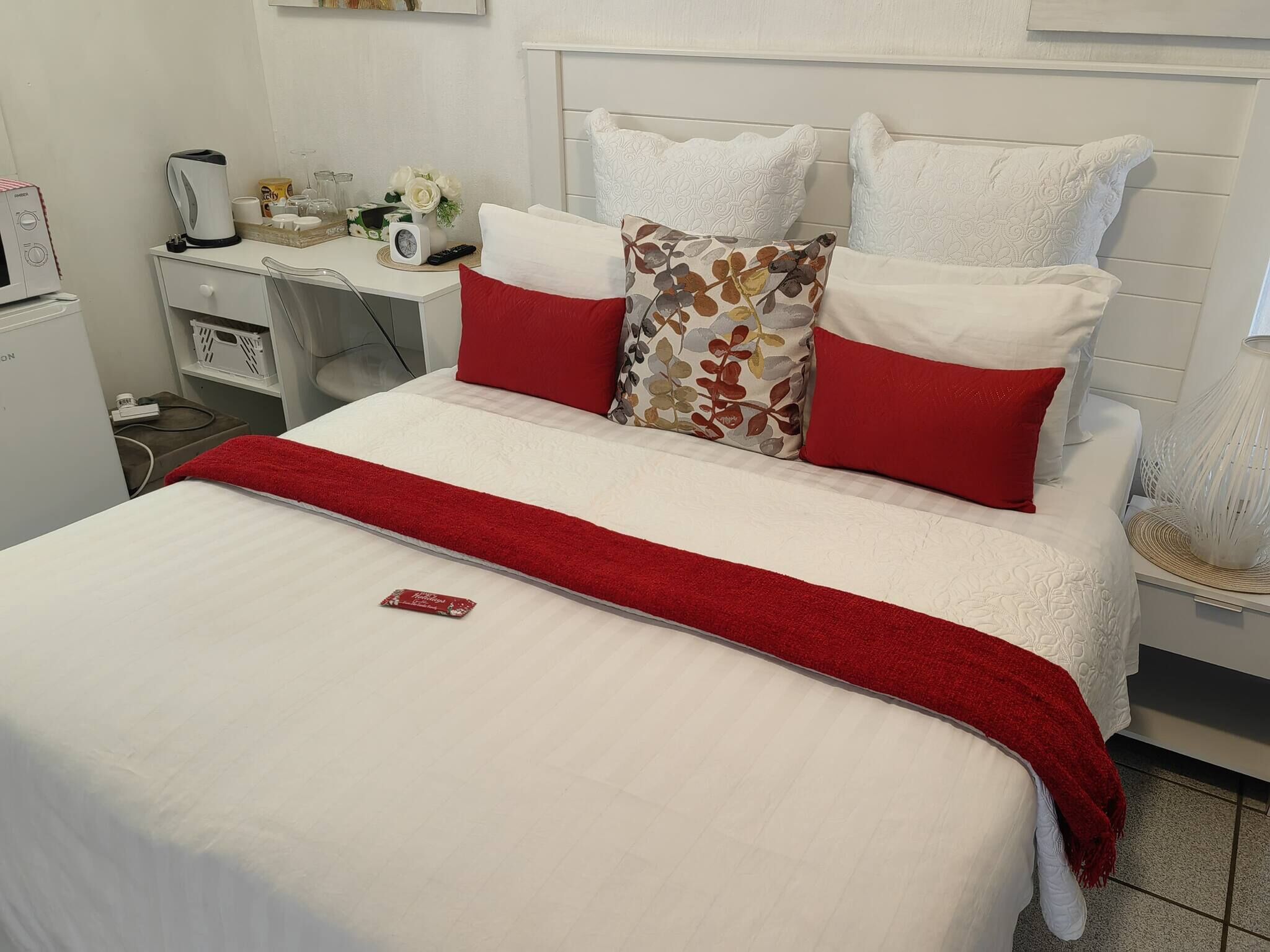A neatly made bed with white and red bedding, a small bedside table with a lamp, and a white wall with a framed picture.