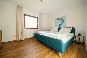 Une chambre avec un plancher en bois, un lit doté d’une tête de lit turquoise, une fenêtre avec des rideaux, un radiateur et une œuvre d’art accrochée au mur.