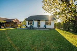 Une maison avec une pelouse bien entretenue, une terrasse avec des chaises et un grand arbre qui offre de l’ombre.