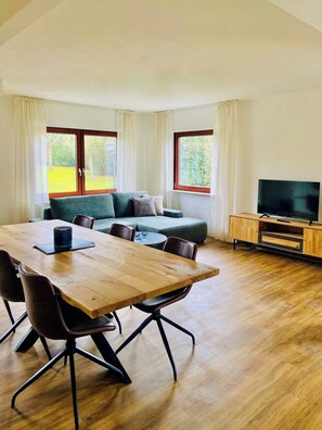 Ein Zimmer mit einem Holzespeisetisch, Stühlen, einer Couch, einem Fernseher auf einem Holztischchen und großen Fenstern mit Vorhängen.