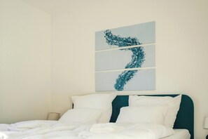 Ein Hotelzimmer mit Bett, weißer Bettwäsche, einem türkisblauen Kopfteil und einem an der Wand befestigten abstrakten Kunstwerk.