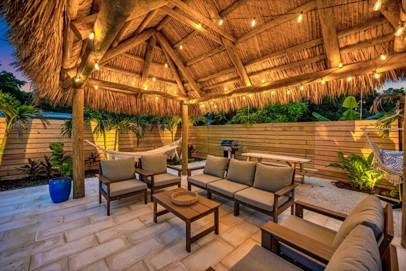 An outdoor patio with wooden furniture, a thatched roof, and string lights.