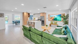 A modern living room with a green sofa, a dining area, a kitchen, and a large TV on the wall. - 2 Resort Homes Side-by-Side - Pool, Tiki, Ice Bath (Plantation)