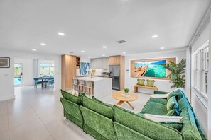 A modern living room with a green sofa, a dining area, a kitchen, and a large TV on the wall. - 2 Resort Homes Side-by-Side - Pool, Tiki, Ice Bath (Plantation)