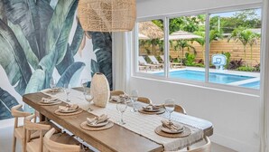 A dining area with a table set for six, a large woven light fixture, and a view of a pool and garden through large windows. - 2 Resort Homes Side-by-Side - Pool, Tiki, Ice Bath (Plantation)