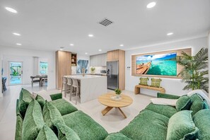 Ein modernes Wohnzimmer mit grünen Sofas, einer zentralen Kücheninsel mit Barhocker, einer Küche mit Kühlschrank und einem Fernseher, der eine tropische Szene zeigt.