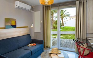 Une pièce avec un canapé bleu, une table ronde sur laquelle se trouvent un chapeau et un livre, un fauteuil rouge et une vue sur un jardin, visible à travers une porte coulissante.