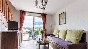 A room with a sofa, a TV on a stand, a table with books and a lamp, and a view of the outdoors. - Lovely Duplex With Scenic Terrace (Carayac)