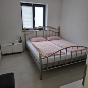Een enkel bed met een metalen frame en hoofdeinde, een nachtkastje met een lamp, en een raam met zonwering.
