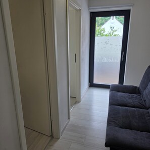 Ein Flur mit einer Tür zu einem Außenbereich, einem grauen Sofa und einem hellen Holzboden.