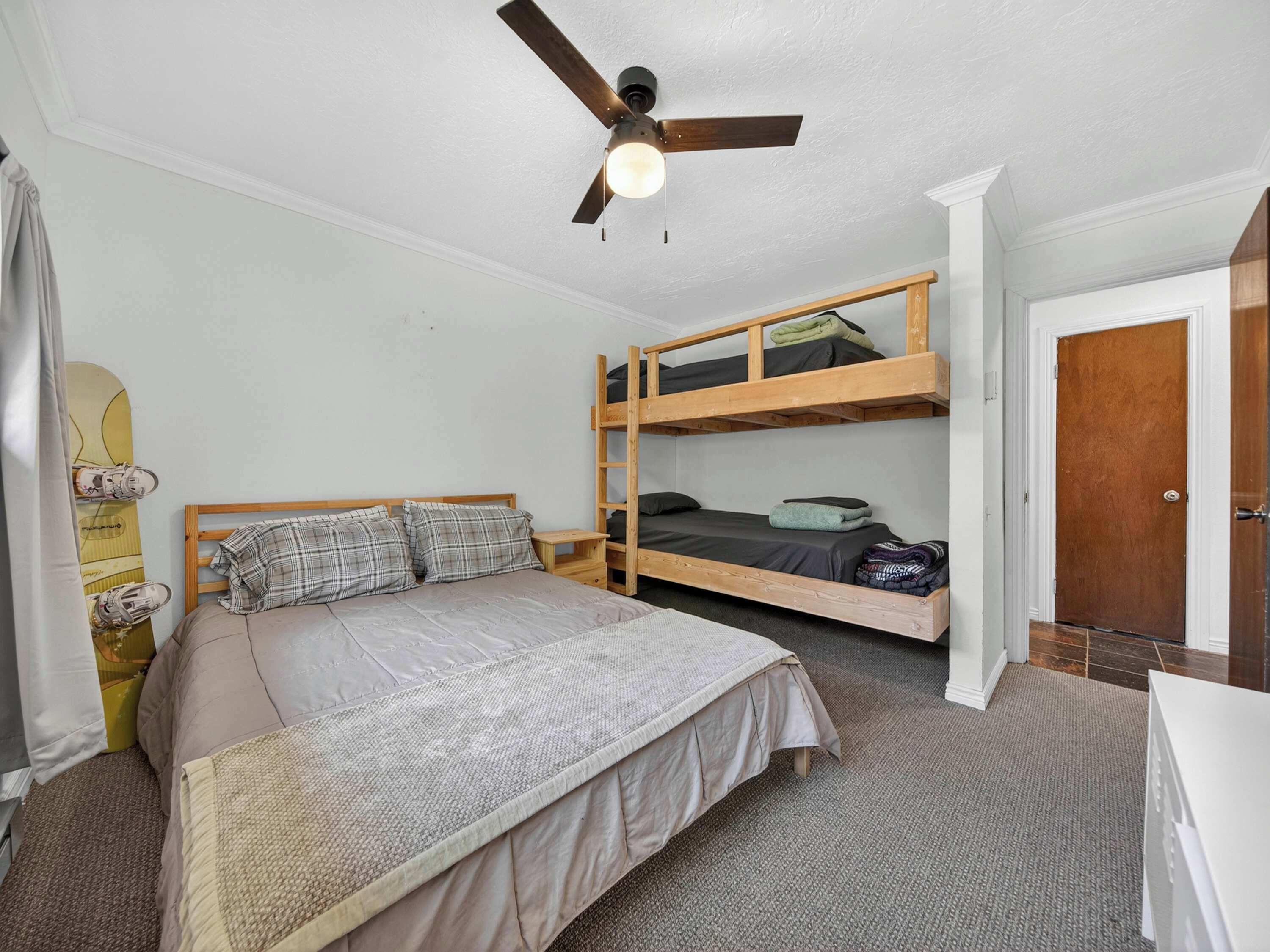 A room with a bunk bed, a single bed, a ceiling fan, a door, and a small table.