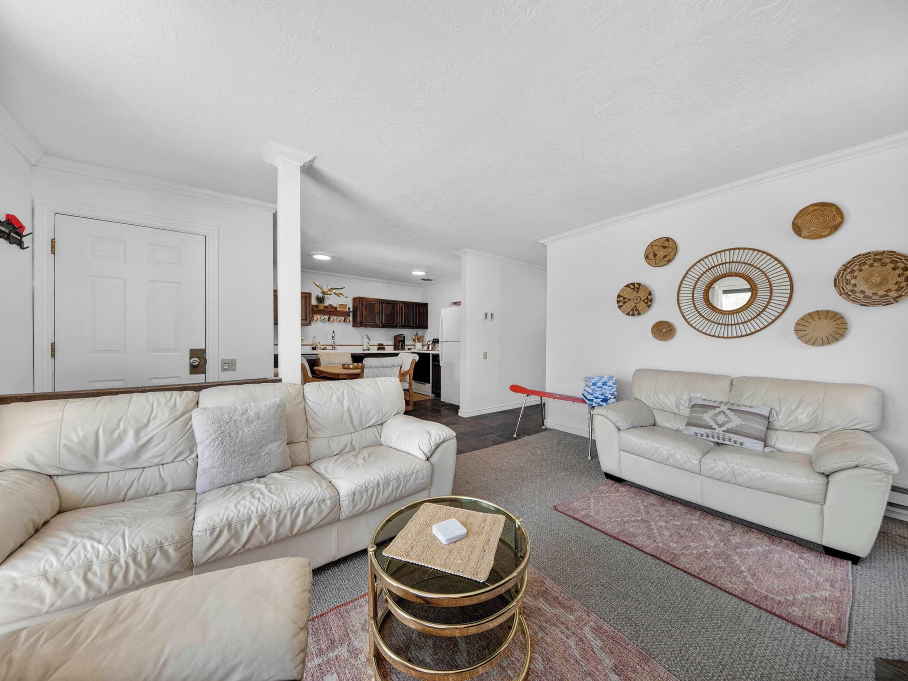 A living room with two white leather sofas, a glass coffee table, and a kitchen in the background.