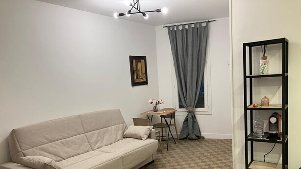 En stue med en sofa, et lille bord med stole, en reol, et vindue med gardiner og en loftlampe.