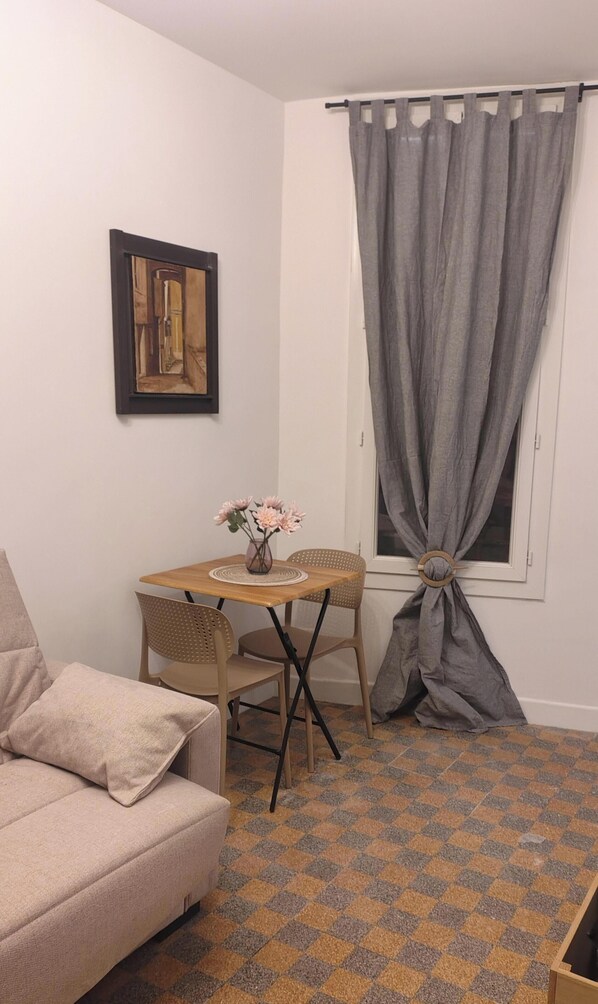 Ein kleines Zimmer mit einer Couch, einem Esstisch mit Stühlen, einem gerahmten Bild an der Wand und einem Fenster mit Vorhängen.