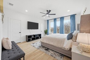 Ein Schlafzimmer mit einem Bett, einem Fernseher, einem Deckenventilator, einer Bank und einem Fenster mit Vorhängen.