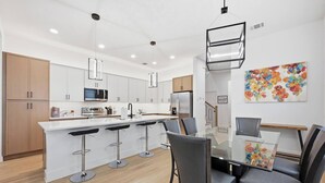 Une cuisine moderne avec des meubles blancs, des appareils en acier inoxydable, un îlot central doté de tabourets, et un coin repas avec une table et des chaises en verre.