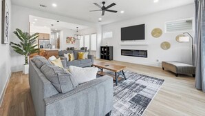 Un salon moderne avec un canapé gris, une table basse, une cheminée, un téléviseur fixé au mur et un ventilateur de plafond.