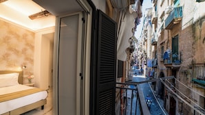 A narrow urban street with buildings on either side, a balcony with a railing, and a bed with a nightstand in the foreground. - NAPOLI TIME TOLEDO (Naples)
