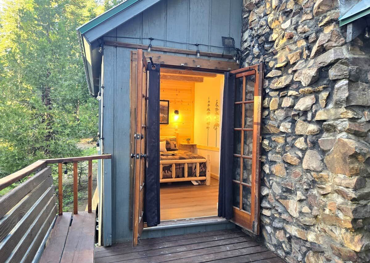 Eine Holzhütte mit Steinfassade, eine offene Tür, durch die ein Bett zu sehen ist, und eine Holterrasse.