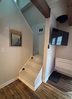 Eine Treppe führt zu einer zweiten Etage, ein an der Wand befestigter Fernseher und ein gerahmtes Poster sind zu sehen.