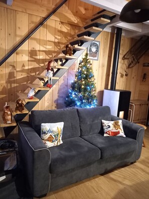 A cozy living room with a dark grey sofa, a wooden staircase, a decorated Christmas tree, and a framed picture on the wall. - Apartment in a chalet in the Haut Jura, ideal for family vacations. (Lamoura)