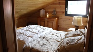 Ein Schlafzimmer mit Holzbalkendecke, einem Bett mit weißer Bettwäsche, einem Nachttisch mit Lampe und einem Fenster mit Jalousien.