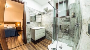 A modern bathroom with a marble wall, a glass shower enclosure, a wooden floor, and a bedside table with a lamp. - Paladini al Casale (Piazza Armerina)
