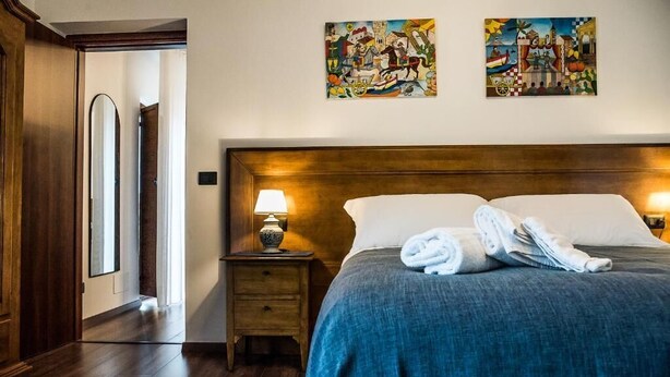 A hotel room with a wooden headboard, a bed with a blue blanket, two paintings on the wall, a nightstand with a lamp, and a door leading to another room. - Paladini al Casale (Piazza Armerina)