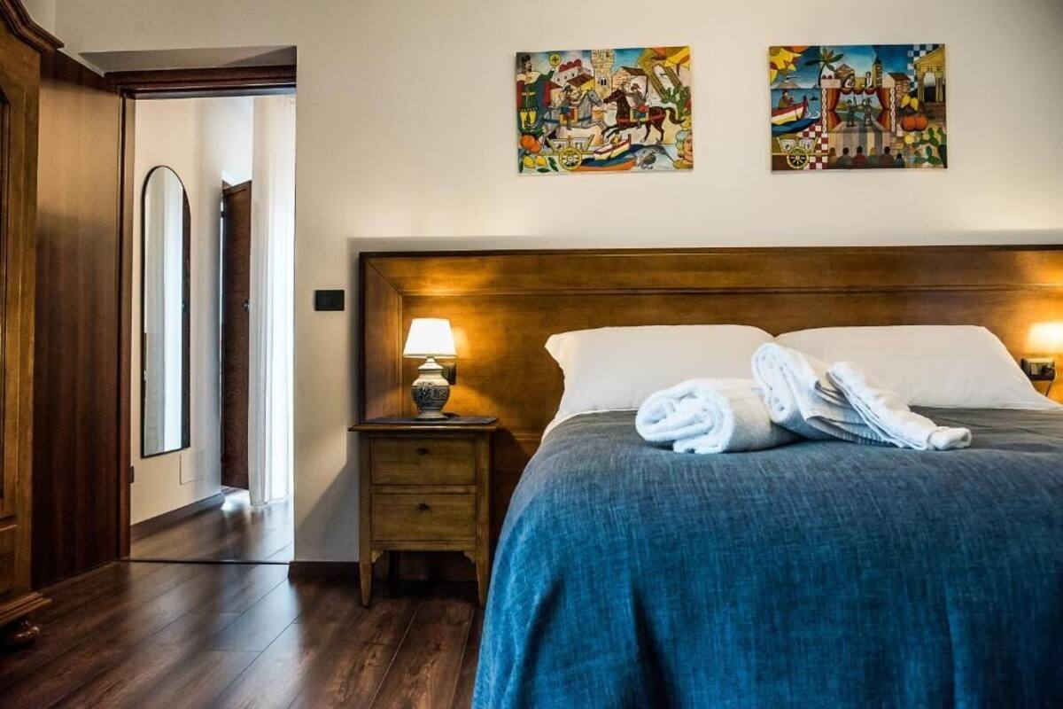 A hotel room with a wooden headboard, a bed with a blue blanket, two paintings on the wall, a nightstand with a lamp, and a door leading to another room.