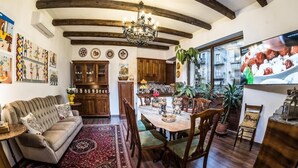 A cozy living room with a sofa, dining table, and chairs, wooden beams on the ceiling, and a kitchen area with cabinets and a television. - Paladini al Casale (Piazza Armerina)