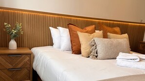 Una cama bien hecha con ropa de cama blanca, una mesita de noche de madera con cajones y una mesita auxiliar con un jarrón de flores.