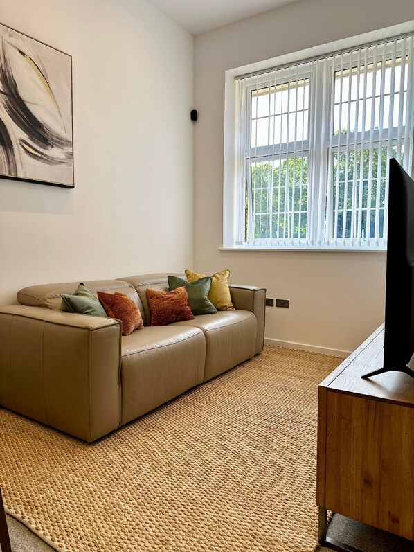 A living room with a beige sofa, a woven rug, a TV stand, and a window with blinds.