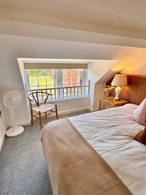 Une chambre à coucher comprenant un lit, une chaise, une table de chevet avec une lampe, une fenêtre et un ventilateur sur pied.