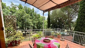Un patio con un tavolo ricoperto da una tovaglia verde e rosa, circondato da piante in vaso e un ringhiera di metallo bianco.