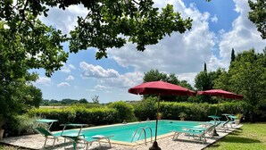 Una piscina con lettini e ombrelloni rossi, immersa nel verde e sotto un cielo sereno.