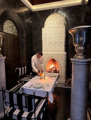 A dining area with a fireplace, a table set for two, and a black and white striped bench. - The riad saeed : A luxury riad in the heart of Marrakech Medina (Marrakesh)