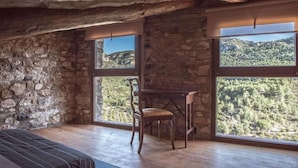 A room with stone walls, wooden beams, a bed, a chair, and large windows with a mountain view.