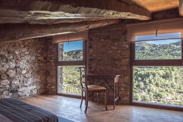 Une pièce aux murs de pierre, avec des poutres en bois, un lit, une chaise et de grandes fenêtres donnant sur la montagne.