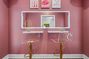 A bathroom with a pink wall, a white vanity with a mirror, and two clear chairs with gold legs. - Bach HQ | Walk to Broadway | Arcade + Pool (Nashville)