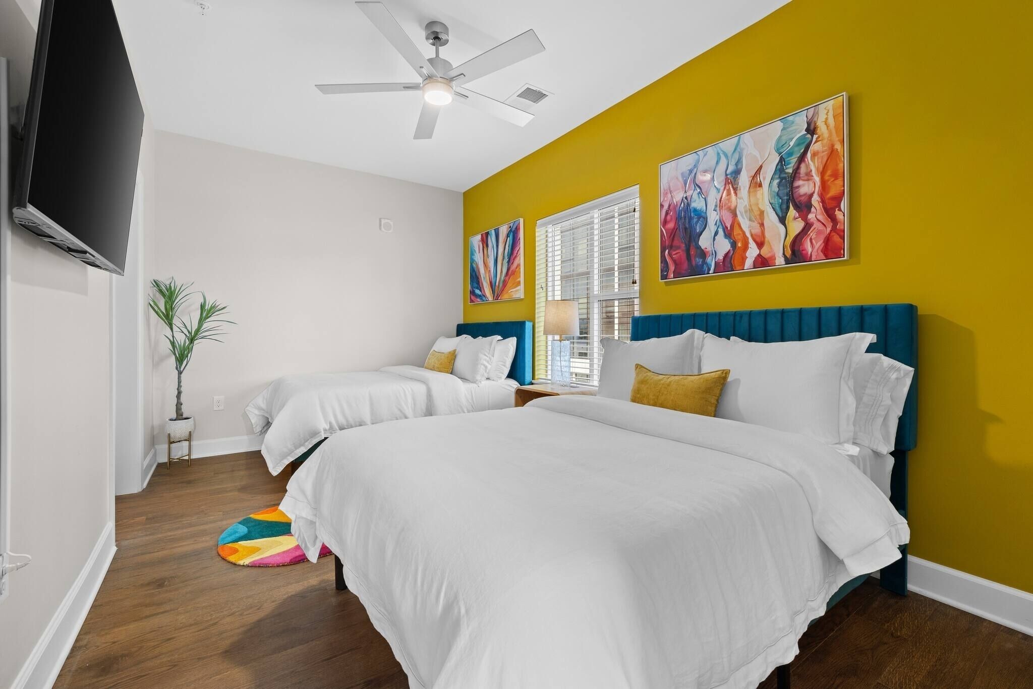 A hotel room with two beds, a ceiling fan, a TV, and artwork on the walls.