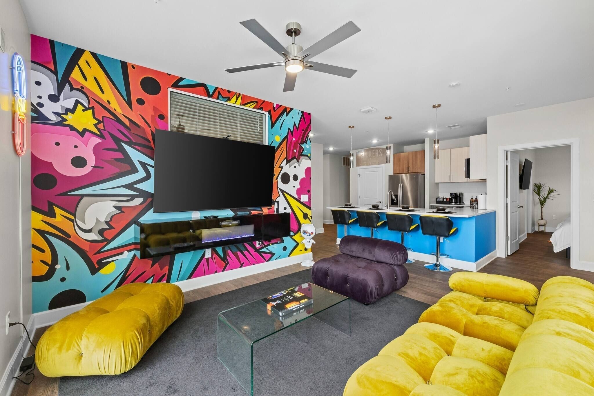 Un salon moderne doté d’un téléviseur à écran plat, d’œuvres d’art aux couleurs vives, d’un ventilateur de plafond et d’un canapé jaune.