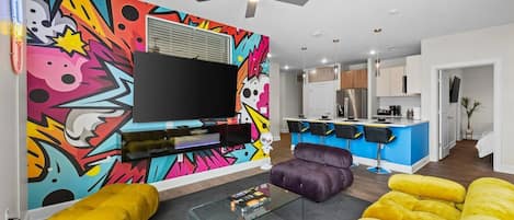 Un salon moderne doté d’un téléviseur à écran plat, d’œuvres d’art aux couleurs vives, d’un ventilateur de plafond et d’un canapé jaune.