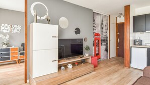 Een moderne woonkamer met een televisie, houten kasten, een kunstwerk van een rode telefooncel en een keukenhoek.