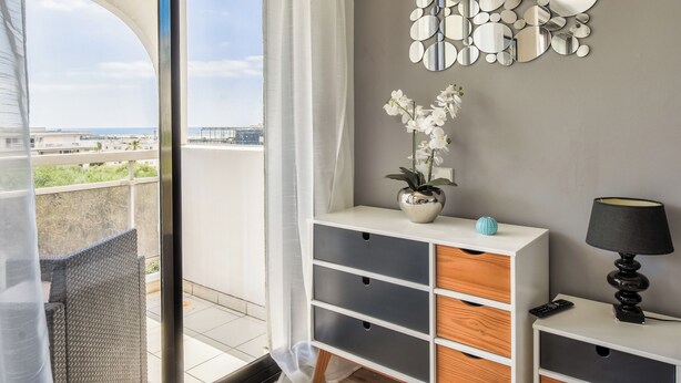 Een moderne kamer met balkon, een spiegelkunstwerk aan de muur, een vaas met witte bloemen, een lamp en een afstandsbediening op een console tafel.