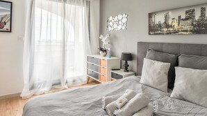 Een slaapkamer met een bed, een nachtkastje, een lamp, een stadslandschap schilderij en een raam met dunne gordijnen.