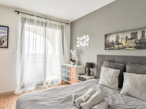A bedroom with a bed, a nightstand, a lamp, a cityscape painting, and a window with sheer curtains. - Apartment with terrace and garage in St-Laurent (Saint-Laurent-du-Var)