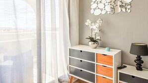 Een kamer met een wit en grijs kastje, een vaas met witte bloemen, een spiegel aan de muur en een lamp.