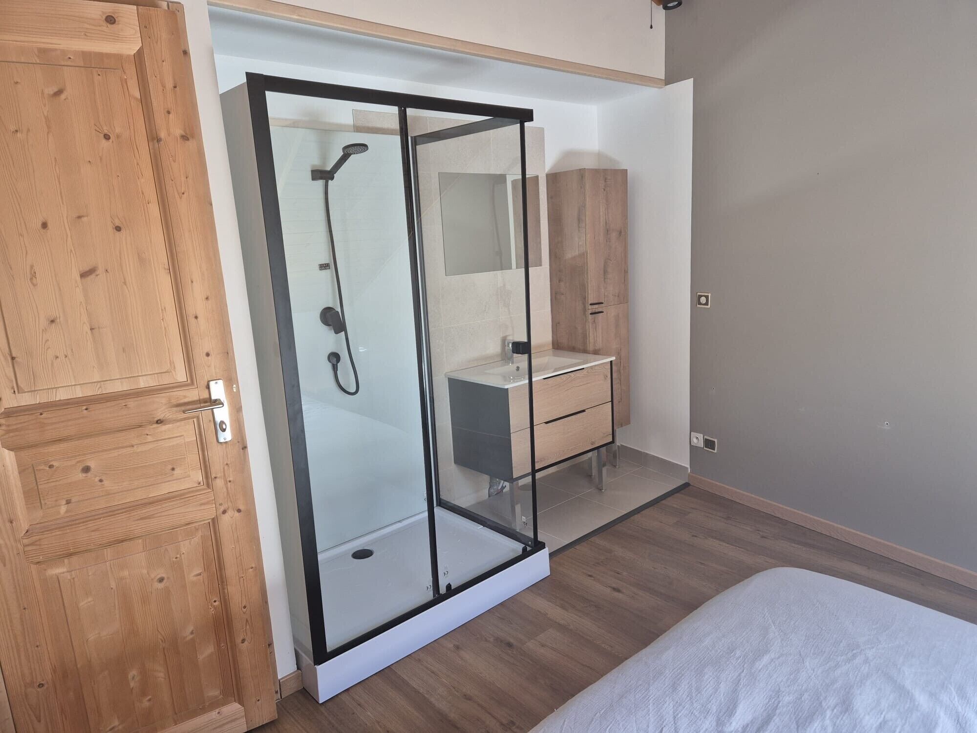 A bedroom with a wooden wardrobe, a glass shower enclosure, a white bed, and a wooden cabinet.
